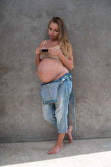 Pretty pregnant woman in denim jumpsuit and bra drinking coffee while standing over gray wall background. Maternity, pregnancy, new life healthy food concept