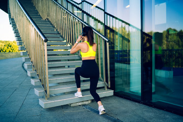 Fototapeta na wymiar Back view of female training lower body muscles outdoors in headphones dressed in tracking wear, slim sports woman with perfect body share squating on stairs listening music from playlist on workout