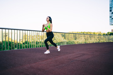 Obraz premium Serious strong female athlete in active trendy wear and headphones training legs and toning outdoors, determined sportswoman listening vitality playlist via sound accessory having morning workout