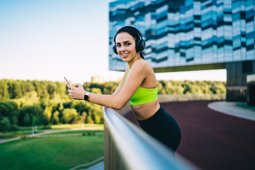 Portrait of cheerful female meloman keeping healthy lifestyle and listening music in headphones and smartphone, happy 20s sports woman enjoying break on training checking playlist on mobile phone