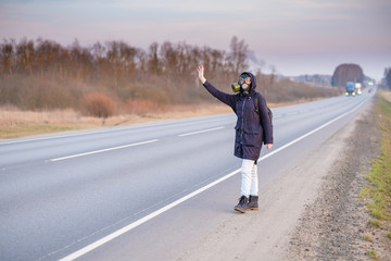 COVID-19. A girl in dark clothes, a gas mask and a hood on her head. Cloudy day in the country. She raises her hand and tries to stop the passing car. Escaping from a zone infected with a coronavirus.