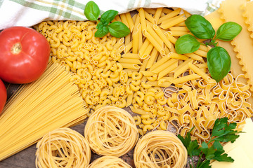 Background composition of different types of raw pasta, with basil leaves and fresh tomatoes.