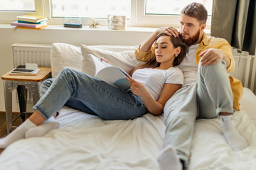 young caucasian married couple relax at home, enjoy being together, have rest lying on bed and reading a book. leisure time, weekends or holidays. indoors