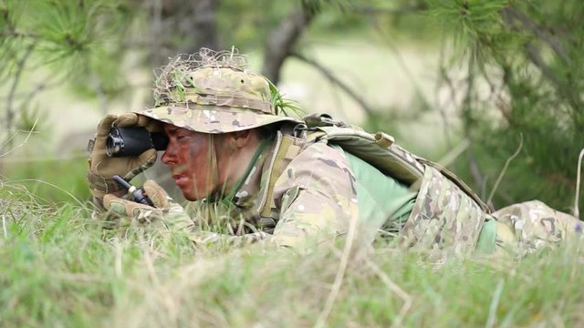 Soldier lies in wait, watching through field glasses, talking on radio.