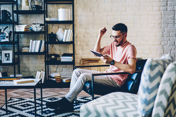 Happy caucasian male gesture raising hand up accomplished next level via digital tablet on free time, hipster guy playing game satisfied with winning high score feeling victorious and using touchpad