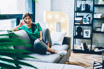 Cheerful caucasian hipster girl satisfied with free time and mobile phone connection talking on mobile phone sitting on couch at living room, smiling woman having cellular conversation on free time