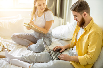 young caucasian married couple sit working at home, man use laptop while his wife taking notes. work environment at home on the bed