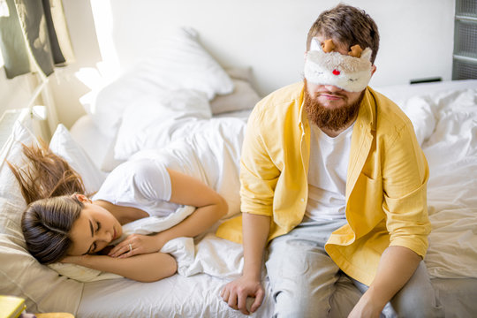 Sleepy Caucasian Man In Mask For Sleeping Sit On Bed Near Sleeping Wife,in The Morning At Home. Man Want To Sleep, Before Going To Work.