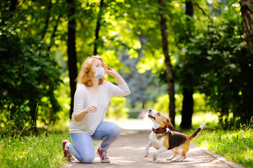 Young beautiful woman wearing disposable medical face mask playing with Beagle dog in the park during coronavirus outbreak. Walking of pets.