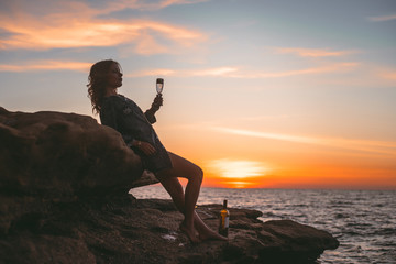 Silhouette of a resting girl on a rock with a glass of wine against the background of the sea.