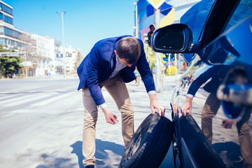 A tired businessman on a sunny day is taking a tire out of his car in order to be able to change his flat tire.