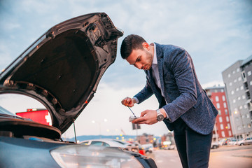 Young manager formally dressed checking his car oil at sunset while leaning under the car's hood.