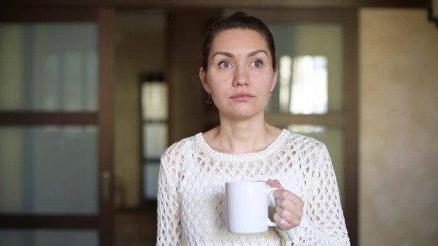 The girl pours red wine into a coffee mug and drinks, looking out the window in the kitchen. A joke outplaying female alcoholism and boredom
