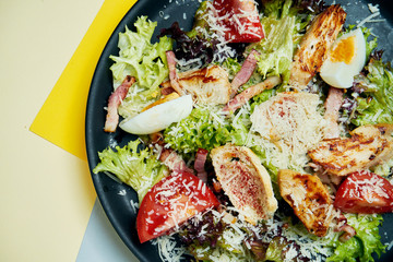 Caesar salad with croutons, parmesan, chicken, egg in black plate on colored background. Restaurant serving. Close up