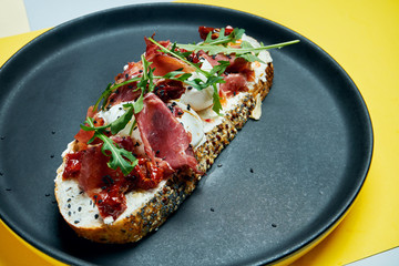 Trendy street snack. Tasty sandwich with jamon, poached eggs, cucumber and microgreen on black plate on colored background. copy space.