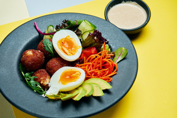 Buddha bowl with chicken croquettes, avcoado, egg, carrot, lettuce and cucumbers in a black plate on a colored background. Close up view. Healthy food
