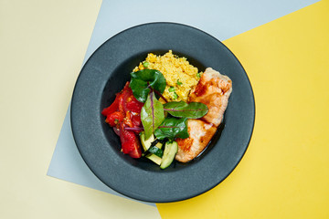 Buddha bowl with salmon filet, dried tomatoes, spinach, cucumber and quinoa in a black plate on a colored background. Close up. Suoer food
