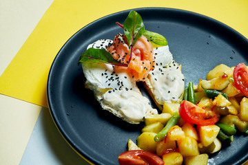 Classic continental breakfast - toasts with cream cheese, salmon and poached egg and a side dish of baked potatoes with tomatoes on a black plate on colored backgrounds