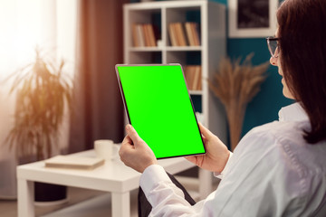 Over shoulder view of woman holding vertical tablet computer with green screen sitting in apartment