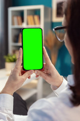 Woman hands vertically holding cellphone with green screen, homey atmosphere