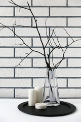 Dry tree branches in a vase on a black tray with candles against a white brick wall