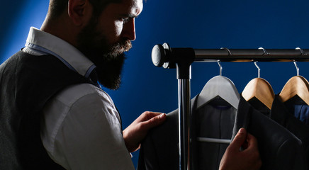 Luxury mens classic suits on rack in elegant men's boutique. Tailor, tailoring. Men's suit, tailor in his workshop. Elegant man's suits hanging in a row. Handsome bearded businessman in classic suit
