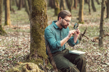Concentrated botanist studying moss through magnifier holding it with tweezers sitting in forest - obrazy, fototapety, plakaty