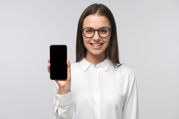Naklejka premium Young business woman wearing formal office shirt and big eyeglasses holding phone with blank screen and presenting it to viewer, copy space, isolated on gray background