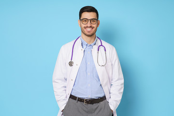 Portrait of smiling young male doctor standing with hands in pockets, isolated on blue background
