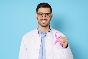 Portrait of young oncologist doctor holding pink ribbon in hand, wearing white coat, isolated on studio blue background. Breast cancer awareness month concept - obrazy, fototapety, plakaty