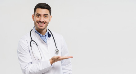 Horizontal banner of smiling young male doctor showing and presenting something with hand, isolated on gray background with copy space on right side