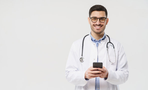 Horizontal banner of young male doctor in white coat smiling, holding his smartphone with both hands, using medical app, standing isolated on studio gray background with copy space on the left side