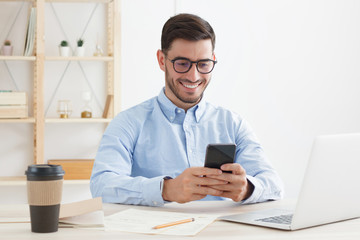 Obraz na płótnie Canvas Young man wearing eyeglasses and shirt in office spending free time scrolling funny posts on his smartphone, smiling and laughing happily while looking at screen