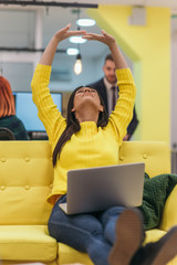 Handsome businesswoman (employee) sitting on a yellow couch while working on her laptop in startup...