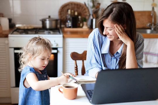 Work From Home Office With Kid. Working Mother Using Laptop. Cozy Freelance Workplace At Kitchen Table. Happy Female Business, Woman Career. Cute Child Drawing In Mom Notebook. Lifestyle Family Moment