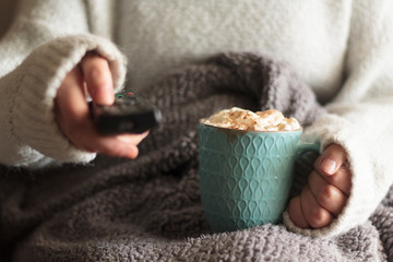 Fototapeta na wymiar Woman covered with blanket holding tv remote and mug of hot drink with whipped cream