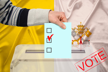 male voter drops a ballot in a transparent ballot box on the background of the vatican national flag, concept of state elections, referendum
