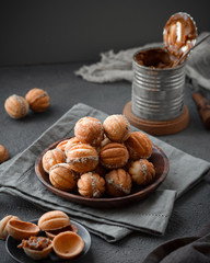 homemade cookies in the form of nuts with a filling of boiled condensed milk stacked in a pyramid in a wooden plate next to a dessert spoon, saucer and a Bank of boiled condensed milk on a gray backgr