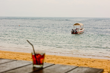 A glass of whiskey with ice on a wooden table in the bar on the sandy shore of the ocean with a view of the waves and the boat.