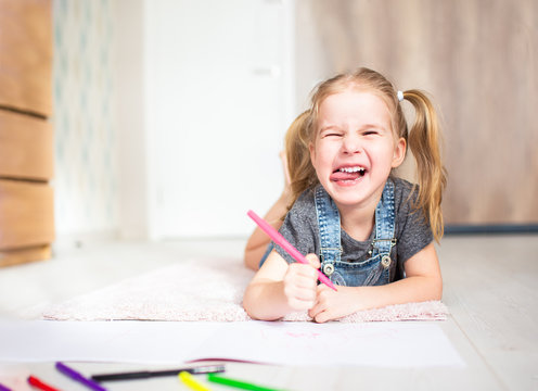Blonde happy little girl with two ponytales drawing and writing lying on the floor at home and showing her tongue. Preschool education, early learning