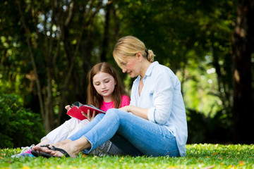 Naklejka na ściany i meble beautyful Mother and cute daughter are reading a book and smile at the park . Happiness and harmony in family life concept.