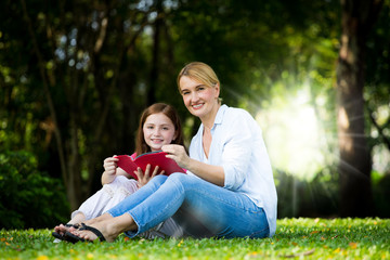 Naklejka na ściany i meble beautyful Mother and cute daughter are reading a book and smile at the park . Happiness and harmony in family life concept.