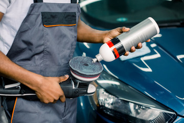 Cropped image of African man, auto service worker, wearing white t-shirt and gray overalls, putting special polish wax or cream on the orbital polisher to polish the blue car behind