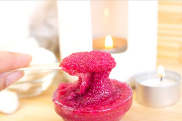 Natural pink body scrub made from sugar. Pink scrub in a glass cup on a background of candles and sugar. stick with a scrub in hand