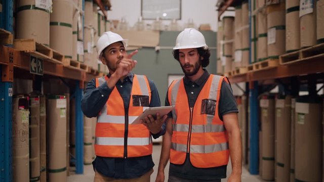 Two warehouse workers wearing hardhats and reflective jackets maintain the detail of manufactured goods in the warehouse using digital tablet