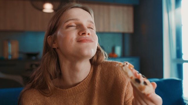 Close up portrait of happy hungry pretty blondehair girl sitting on blue sofa at home and eating tasty pizza, enjoying and smiling