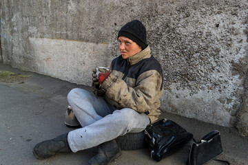Homeless dirty woman sits under the wall and drinks tea from a mug