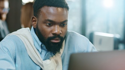 Obraz premium Portrait african american young handsome male uses laptop for work in modern office programmer serious looks at monitor screen typing