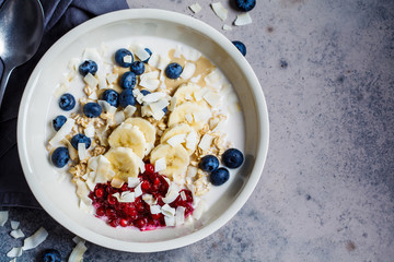 Overnight oatmeal with berries, banana, jam and coconut in a white bowl, top view, gray background.