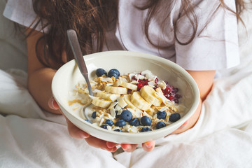 Overnight oatmeal with berries, banana, jam and coconut in a white bowl in hands.